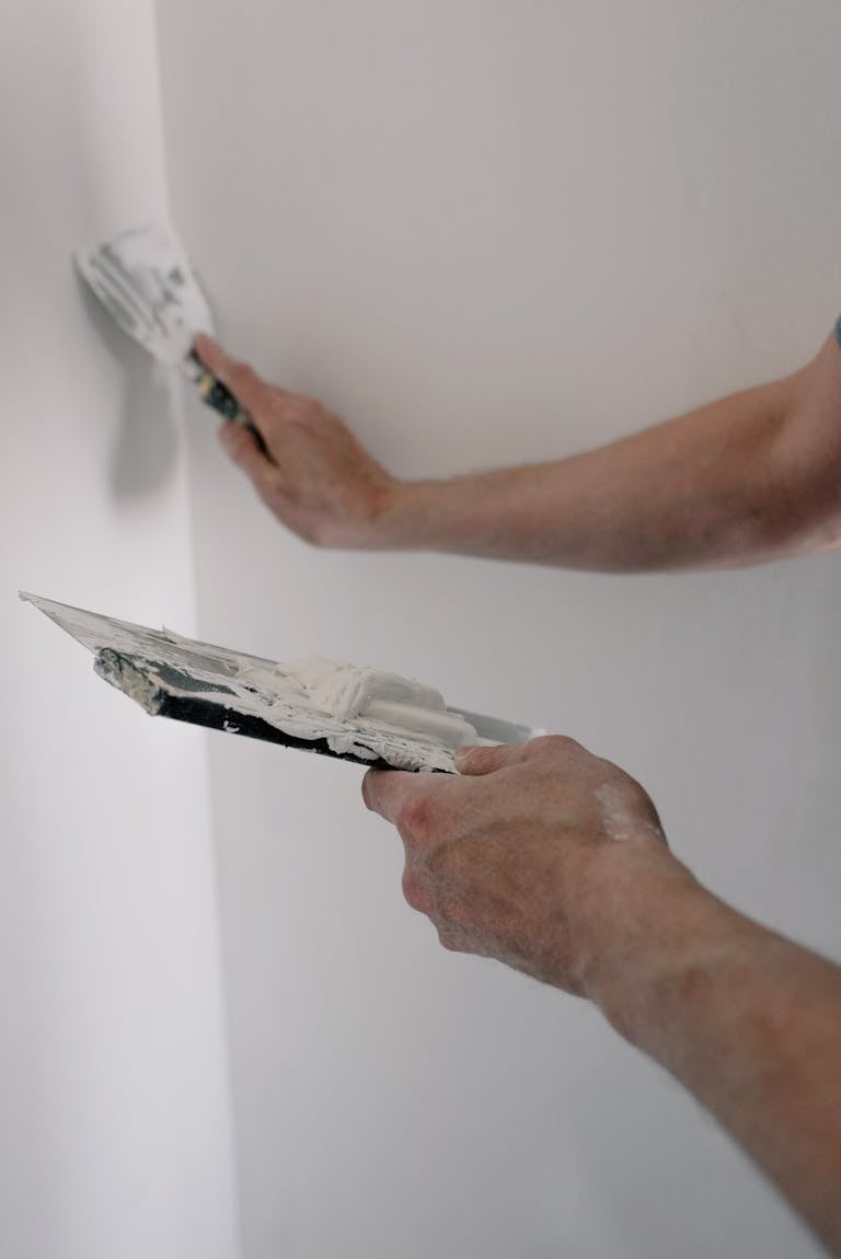 Hands apply spackling paste on a wall using tools, capturing a DIY renovation scene.