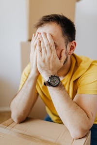A stressed man sits with his hands covering his face amid moving boxes, depicting moving stress.