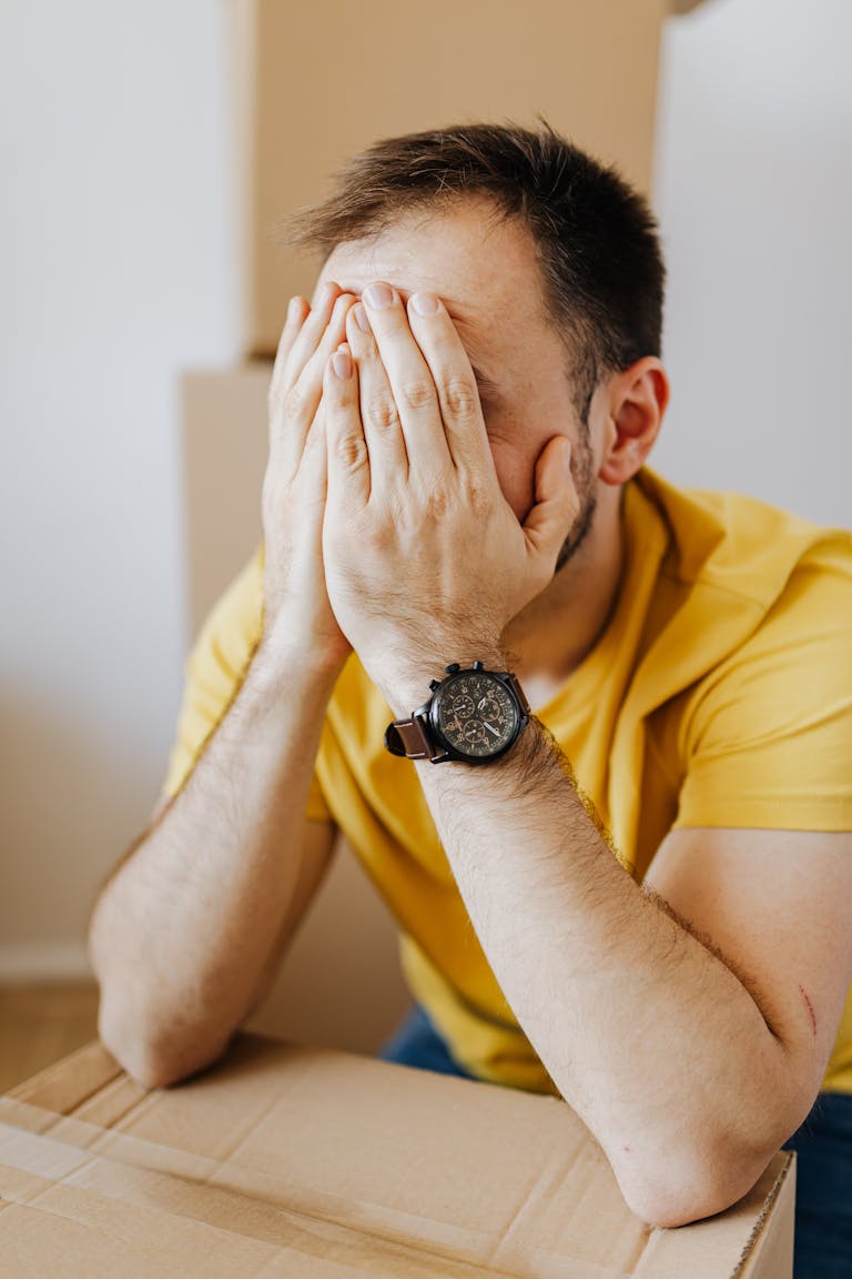 A stressed man sits with his hands covering his face amid moving boxes, depicting moving stress.
